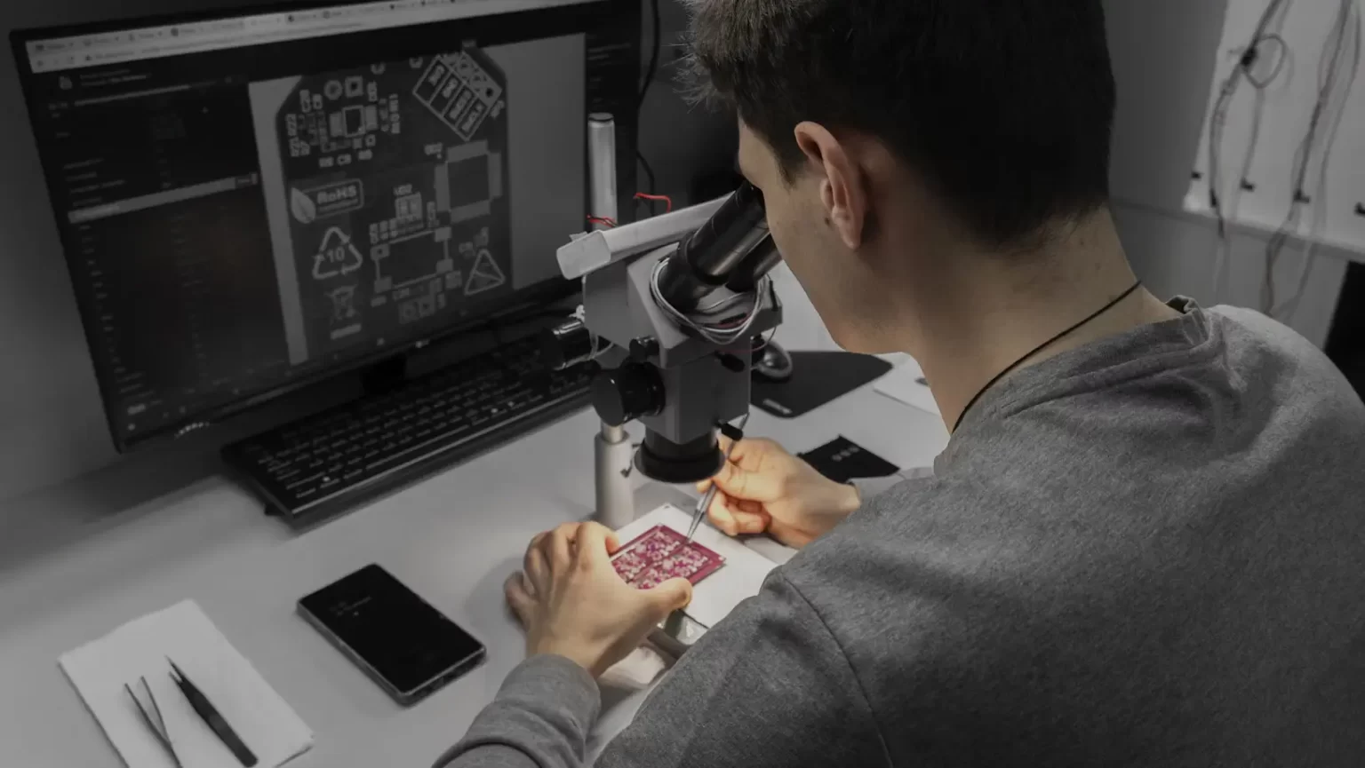 Person examining an electronic circuit board under a microscope, with a computer displaying a circuit schematic in the background.