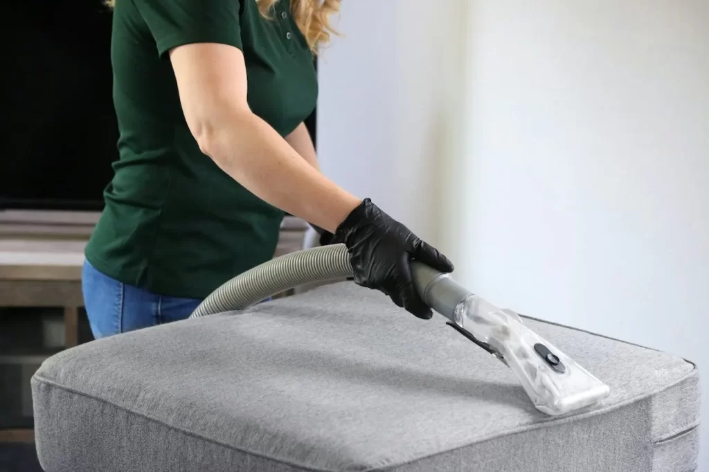 A person cleaning a grey fabric sofa using a modern upholstery vacuum cleaner.
