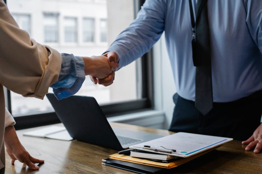 Two professionals shake hands over a desk with a laptop and a stack of files, symbolizing a successful business partnership.
